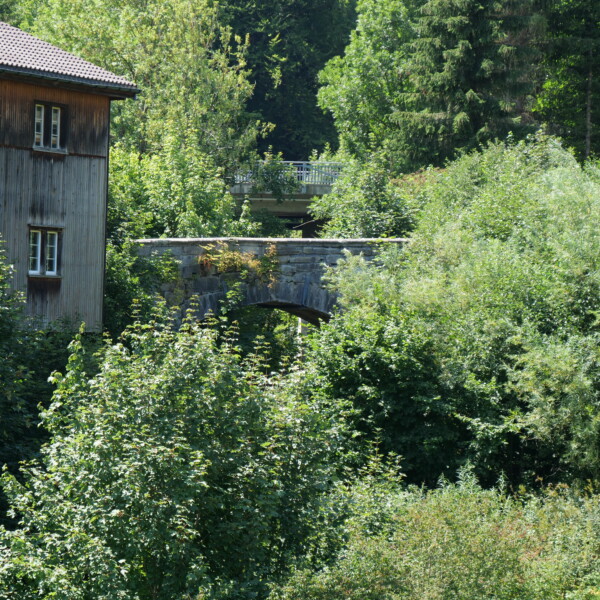 Steinbrücke in Kaubach, umgeben von üppigem Grün und einem Holzhaus.