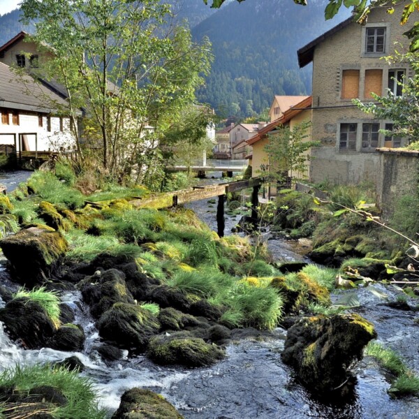 La Noiraigue: Flusslauf mit moosbedeckten Steinen und Häusern im Hintergrund.