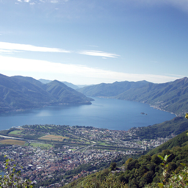 Blick auf Maggia und den Lago Maggiore, umrahmt von Bergen unter blauem Himmel.