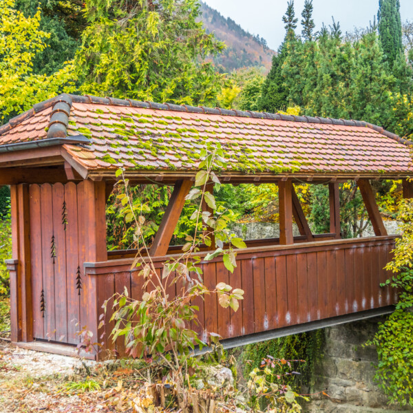 Gedeckte Holzbrücke über den Mümliswiler Bach, umgeben von üppigem Grün.
