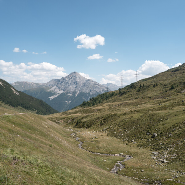 Ova d'Alvra: Berglandschaft mit Wiesen, Bach und schneebedecktem Gipfel unter blauem Himmel.