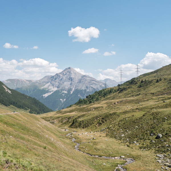 Tal Ova d'Alvra mit Bachlauf, grünen Wiesen und Bergpanorama unter blauem Himmel.