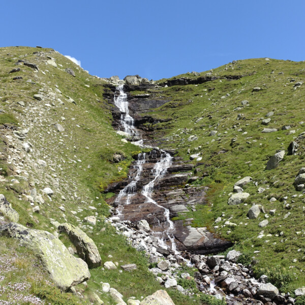 Wasserfall Patthornbach fließt über Felsen in grüner Landschaft unter blauem Himmel.
