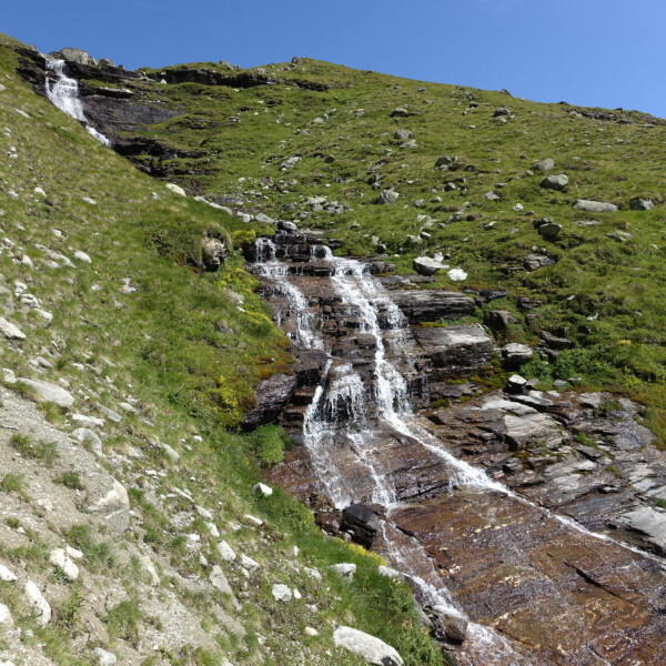 Patthornbach Wasserfall fließt über grüne Hänge und Felsen unter blauem Himmel.