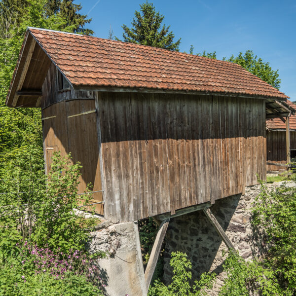 Holzstadel auf Steinfundament in Ranzach, umgeben von Grün. Das Dach ist mit roten Ziegeln gedeckt.
