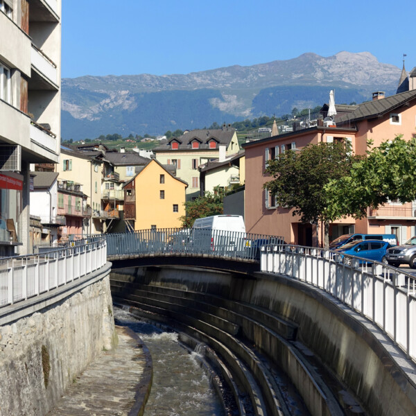 Sionne: Flusslauf durch Sion mit Brücke und Bergpanorama im Hintergrund.