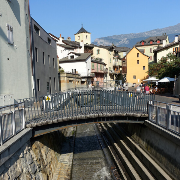 Brücke über den Kanal in Sionne, mit Blick auf malerische Gebäude und Berge.