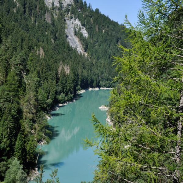 Türkisfarbener Stausee Solis, umgeben von grünen Bäumen und einem bewaldeten Berghang.