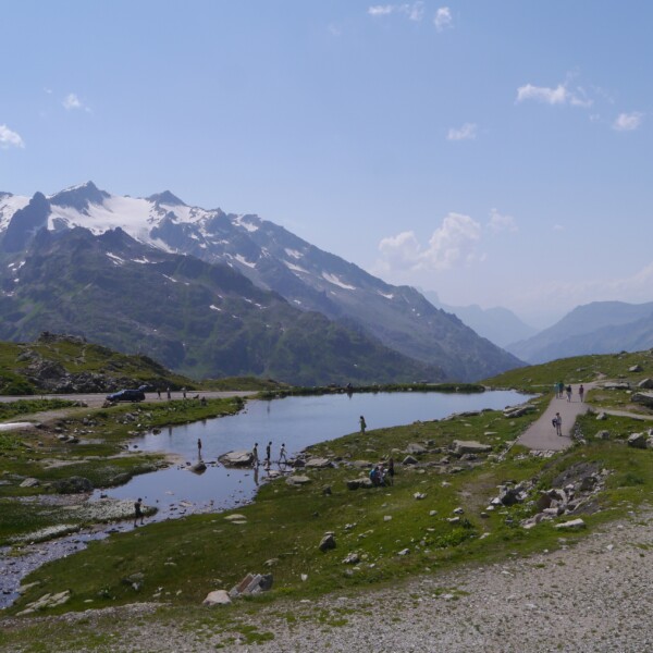 Steinsee mit schneebedeckten Bergen im Hintergrund und Wanderern am Ufer.
