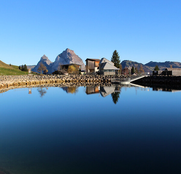 Stoos Seeli: Ruhiger Bergsee mit Spiegelung von Häusern und Bergen unter blauem Himmel.