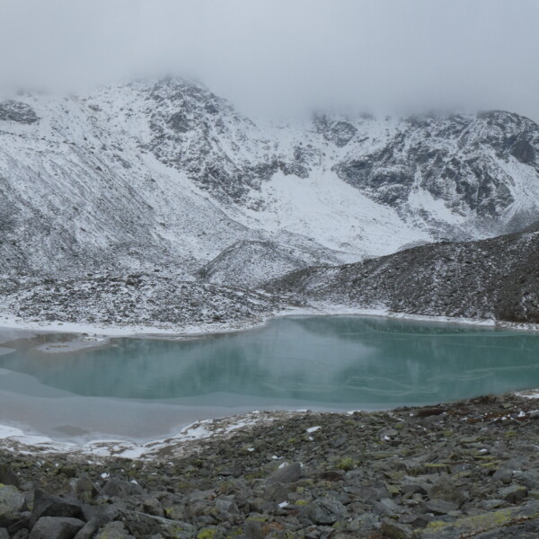 Türkisblauer Tambosee unter schneebedeckten Bergen. Nebel hängt in der Luft.