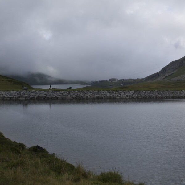 Tannensee-See mit Steinmauer, bewölktem Himmel und grünen Hügeln. Wanderer sind zu sehen.