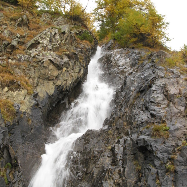 Wasserfall fließt über dunkle Felsen, Bäume im Hintergrund. Tièche.