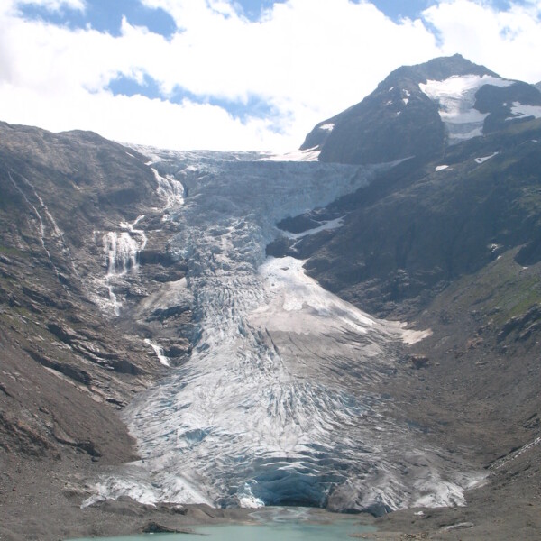 Triftgletscher fließt in den Triftsee, umgeben von Bergen unter blauem Himmel.