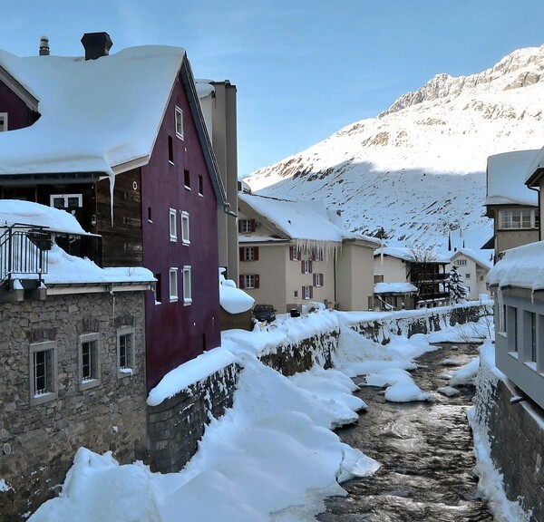 Unteralpreuss im Winter: Verschneite Häuser am Fluss mit Bergen im Hintergrund.