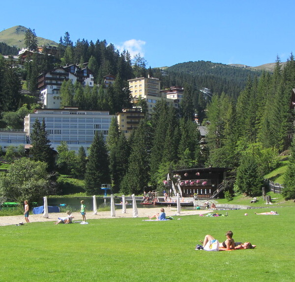 Untersee Arosa: Grünfläche mit Badegästen, umgeben von Bergen und Häusern.