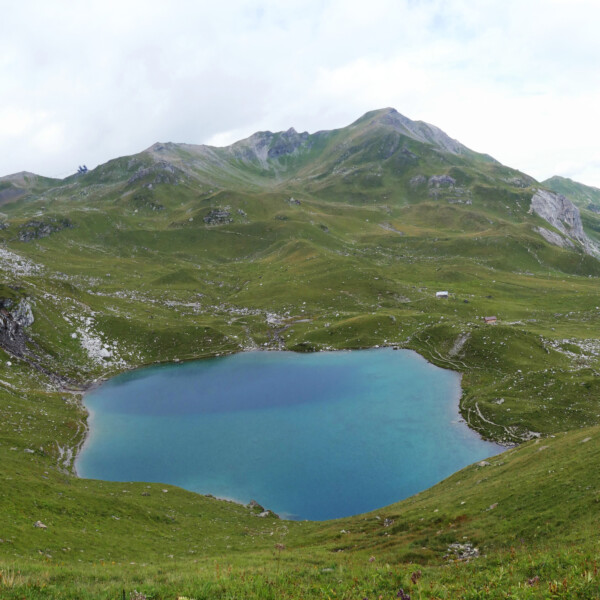 Urdensee, malerischer Bergsee in grüner Alpenlandschaft mit Wanderweg und Gipfeln im Hintergrund.