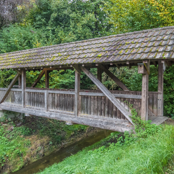 Gedeckte Holzbrücke über einen Bach in Urtenen. Die Brücke hat ein moosbewachsenes Ziegeldach.