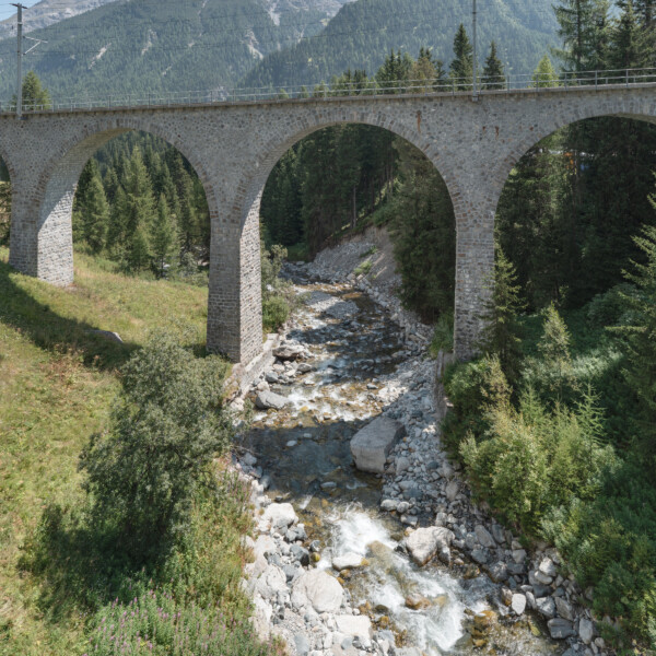 Steinbrücke über den Bach im Vallember mit Bergen im Hintergrund.
