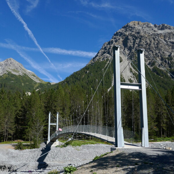 Hängebrücke über den Welschtobelbach, umgeben von Bergen und Wald unter blauem Himmel.