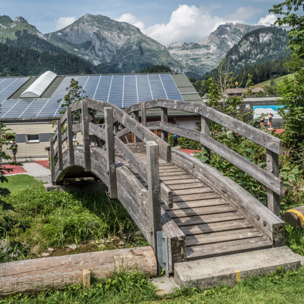 Holzbrücke in Wildhauser Thur mit Bergen und Solarpanelen im Hintergrund.