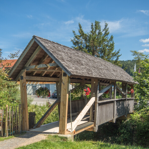Überdachte Holzbrücke in Zwislenbach mit Blumenampeln und traditioneller Bauweise.