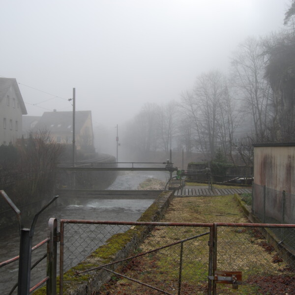 Nebliger Blick auf den Augstbach mit Brücke und Uferbefestigung. Düstere Flusslandschaft.
