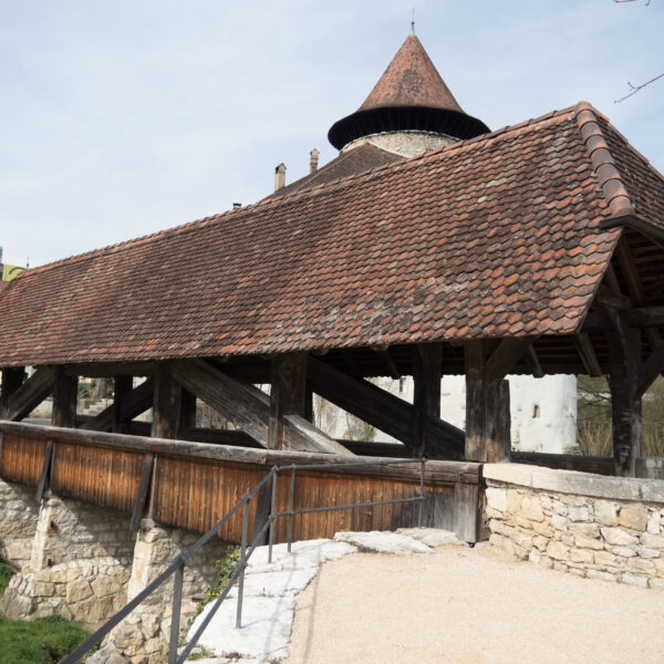 Gedeckte Holzbrücke über die Birs mit Turm im Hintergrund.
