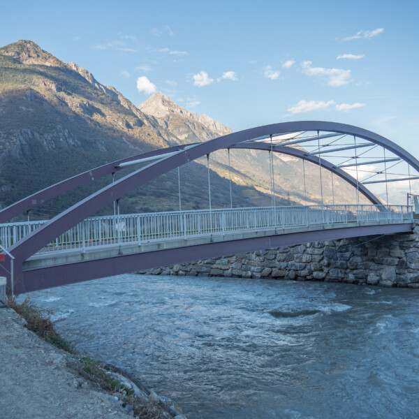 Brücke über die Drance mit Bergen im Hintergrund. Text auf Schild: CAUTION