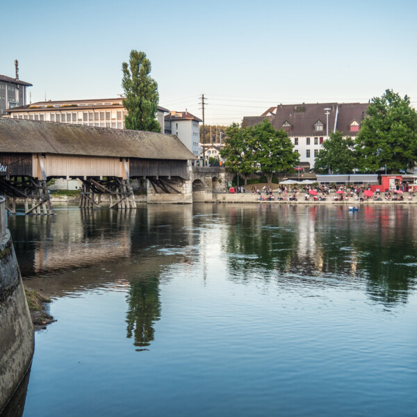 Gedeckte Holzbrücke über die Dünnern mit Badegästen im Hintergrund. 