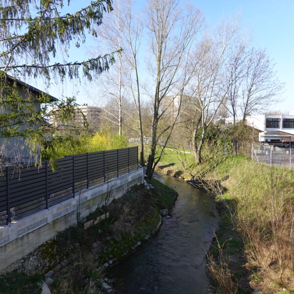 Foron-Flusslauf mit Uferbefestigung, Bäumen und Gebäuden im Hintergrund.