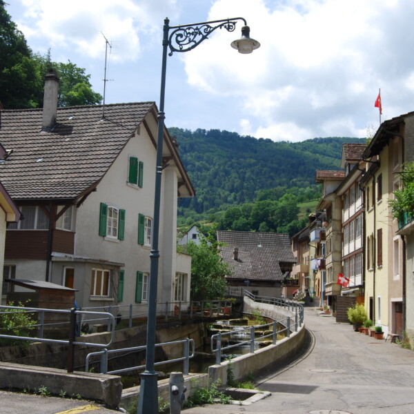 Dorfstraße in Frenke, Schweiz, mit traditionellen Häusern und Schweizer Flaggen.