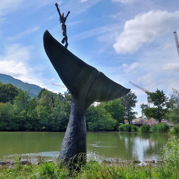 Wal-Skulptur im Giessenparksee mit Figur, Bäumen und Baukränen im Hintergrund.