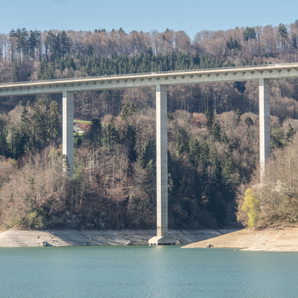 Graue Betonbrücke über den Greyerzersee, umgeben von Bäumen und blauem Wasser.