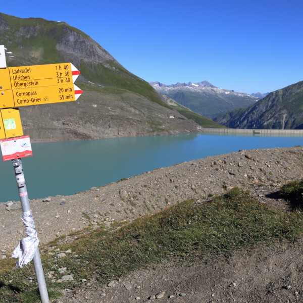 Wanderwegweiser am Griessee mit Blick auf den See und die Berge. Text: Ladstafel, Ulrichen, Cornopass.