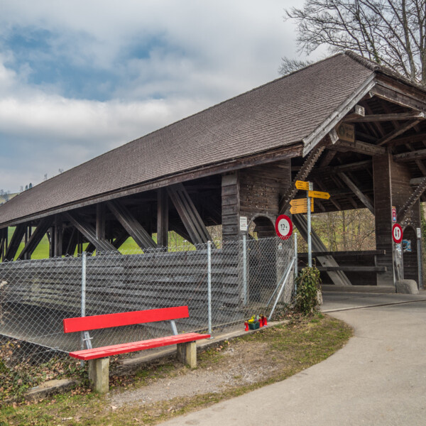 Gedeckte Holzbrücke über die Grosse Melchaa mit roter Bank und Wegweiser.