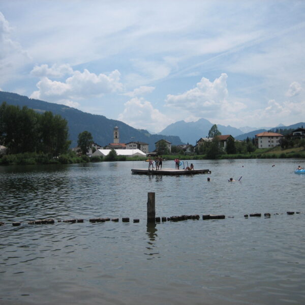 Laaxersee: See mit Badegästen, Bergen und Dorf im Hintergrund.