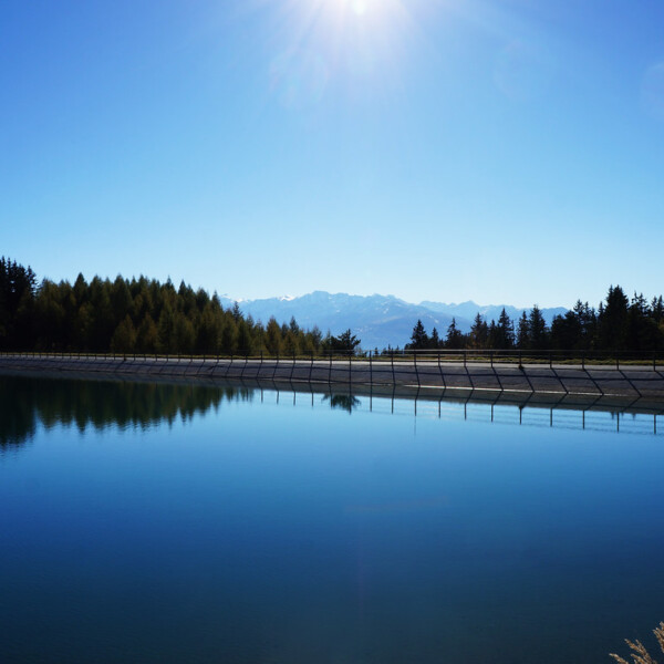 Lac de Chermignon: Ruhiger Bergsee mit Spiegelung von Bäumen und Bergen unter blauem Himmel.