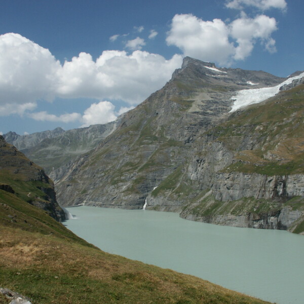 Lac de Mauvoisin: Bergsee mit Gletscher und grünen Hängen unter blauem Himmel mit Wolken.