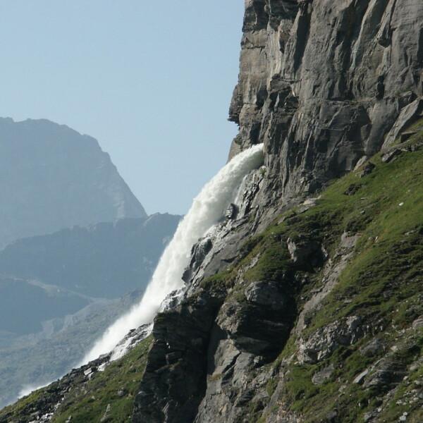 Wasserfall am Lac de Mauvoisin, der von einer steilen Felswand herabstürzt. Grüne Vegetation bedeckt die Hänge.