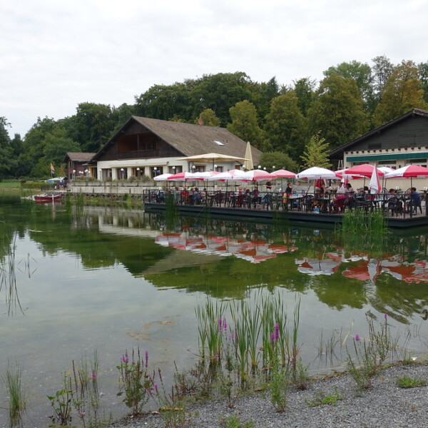 Restaurant am Lac de Sauvabelin mit Gästen unter Sonnenschirmen, Spiegelung im Wasser.