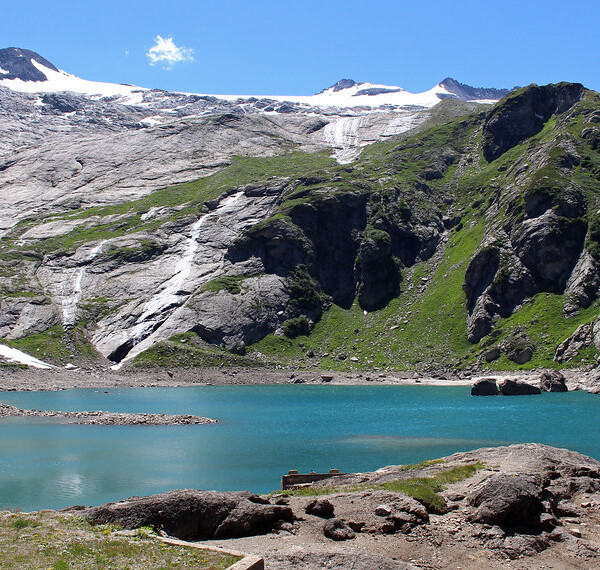 Türkisfarbener Lago del Zött vor schneebedeckten Bergen und grünen Hängen.