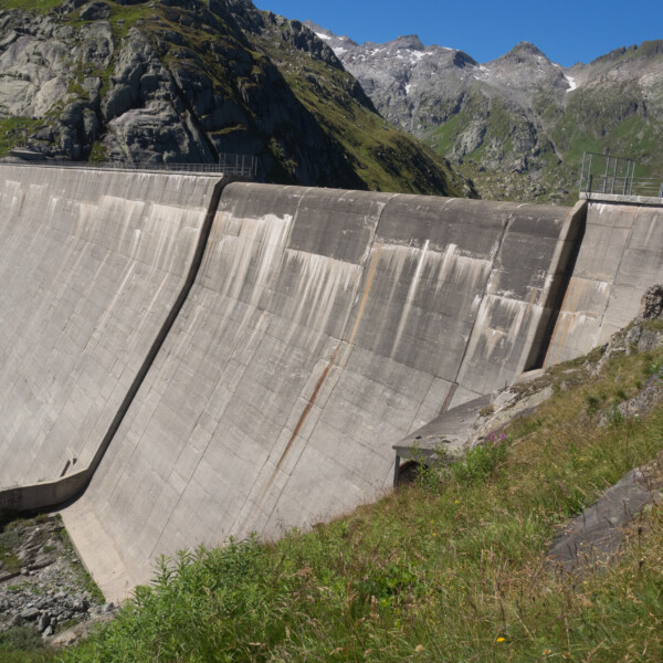 Lago di Lucendro Staumauer vor Bergkulisse. Betonmauer mit Gras im Vordergrund.