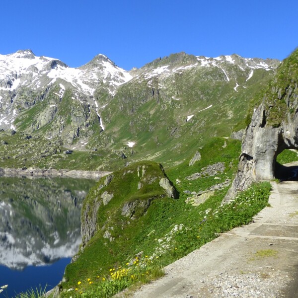 Lago di Lucendro: Bergsee mit Spiegelung, Tunnel im Fels, grüne Landschaft und schneebedeckte Gipfel.