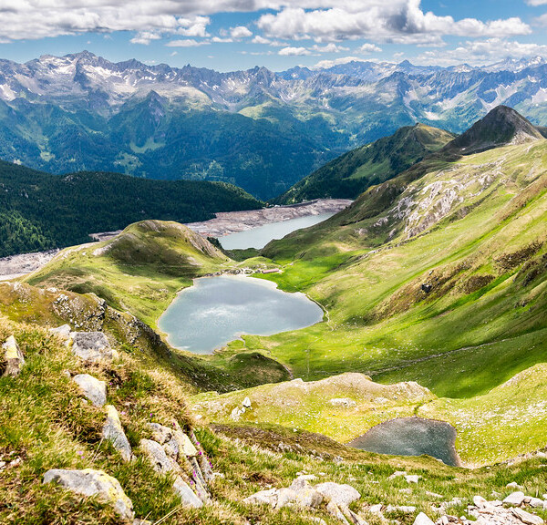 Berglandschaft mit grünen Hängen und dem Lago di Tom, umgeben von schneebedeckten Gipfeln.