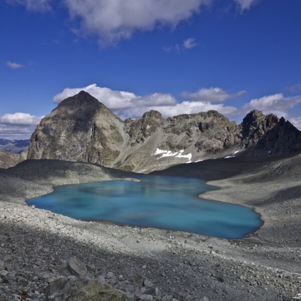 Türkisblauer Bergsee Lej Lagrev vor felsiger Bergkulisse unter blauem Himmel.