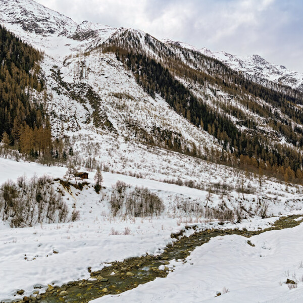 Winterlandschaft mit schneebedeckten Bergen und einem Bach. Lonza im Hintergrund.