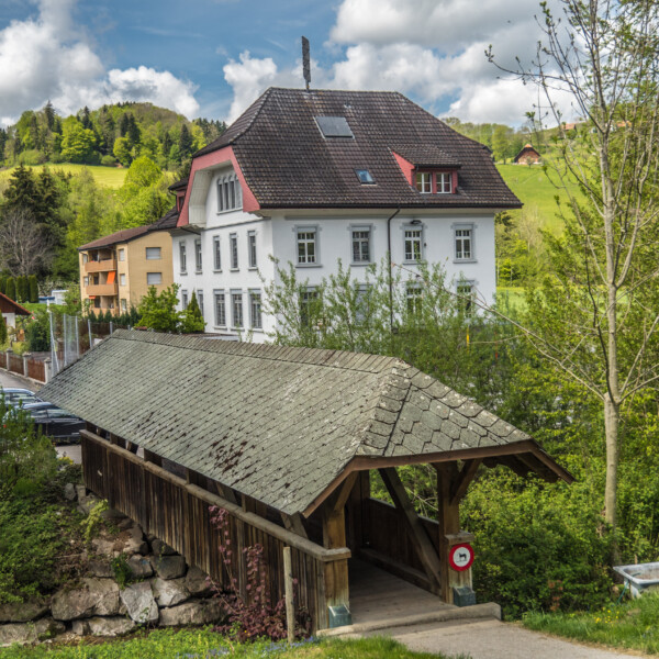 Luthern: Überdachte Holzbrücke mit einem traditionellen Schweizer Haus im Hintergrund.