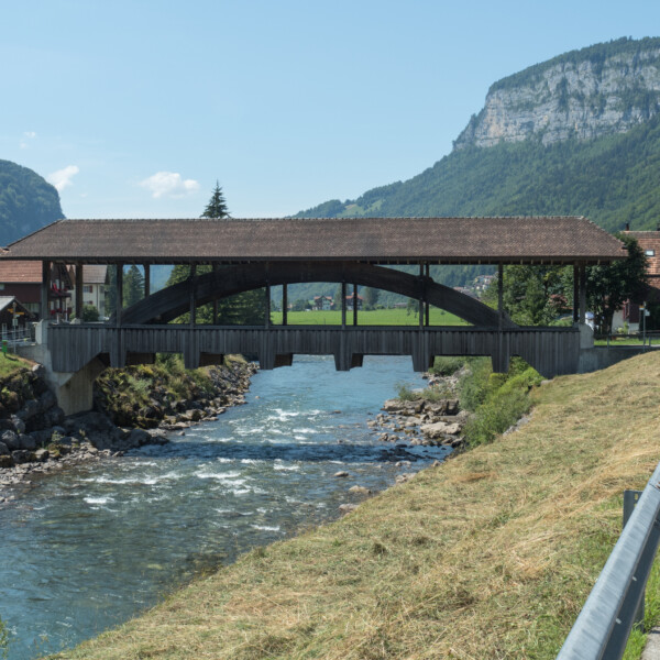 Gedeckte Holzbrücke über den Fluss Muota in der Schweiz vor Bergkulisse.