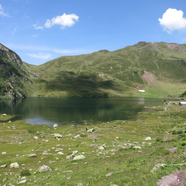Oberer Murgsee: Idyllischer Bergsee mit grünen Hängen und kleinen Hütten unter blauem Himmel.